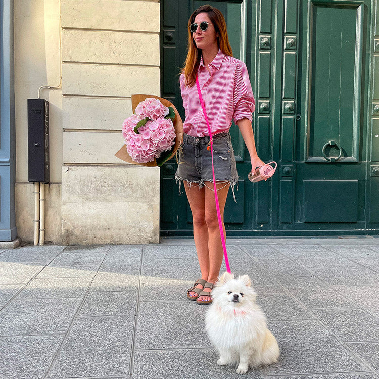 Woman in pink shirt and denim shorts holding flowers and a leash with a small white dog on a city street.