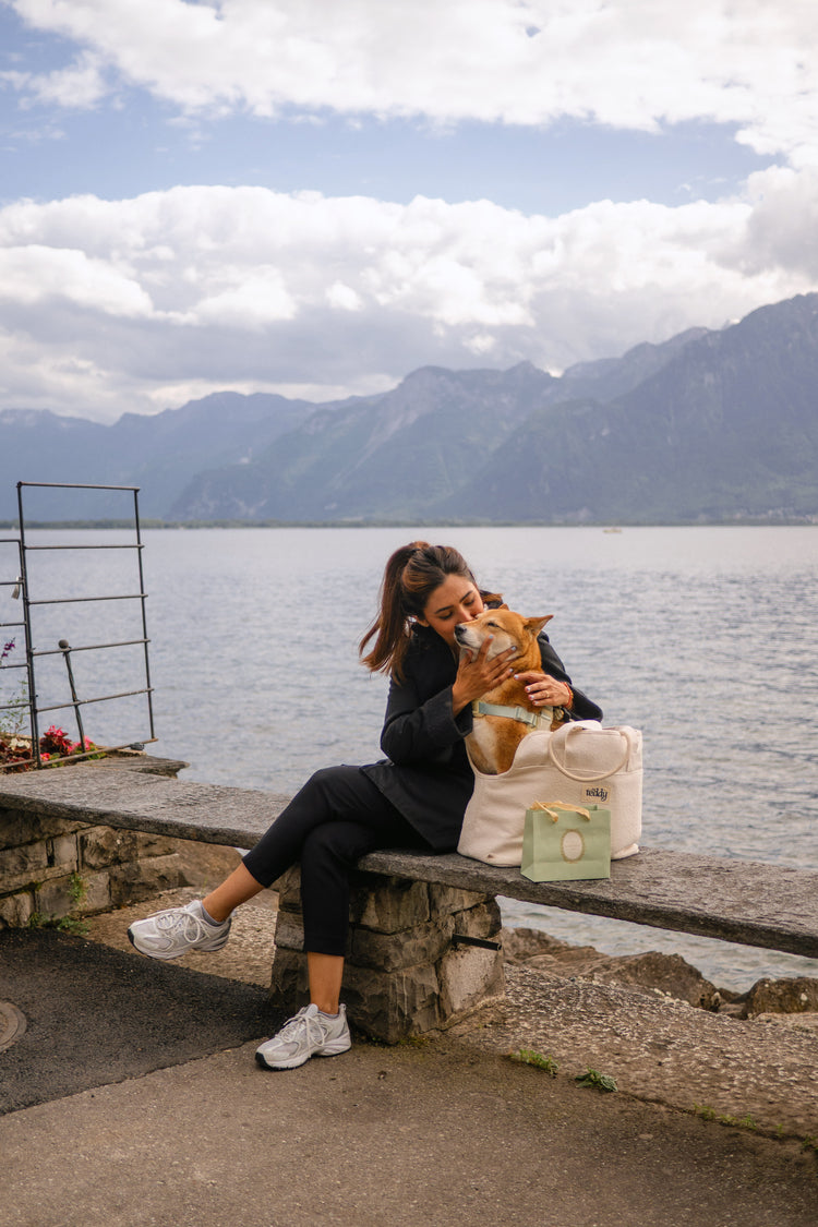 Woman sitting by a lake with mountains in the background, kissing a dog.