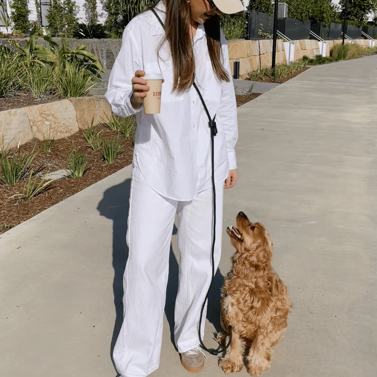 Person in a white outfit with a dog on a leash, holding a coffee cup outdoors.