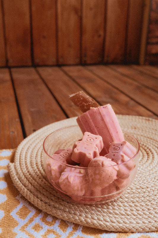 Pink dog ice cream bars in a glass bowl on a woven mat with a wooden floor background
